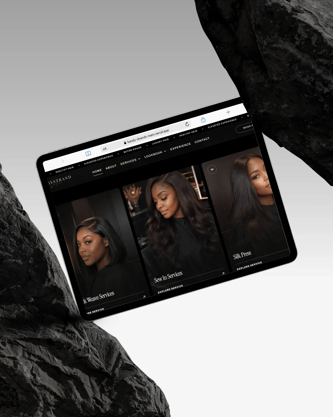 An iPad resting between black rocks, open on the service cards — Quick Weave, Sew In and Silk Press
